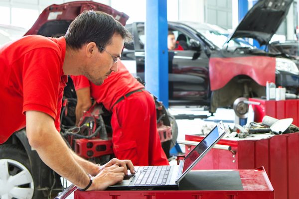 Group of mid aged male car mechanics at work performing regular maintenance. The closest mechanic is using laptop .Wearing red uniforms and standing within individual work spots. Blurry mechanics and cars in background.